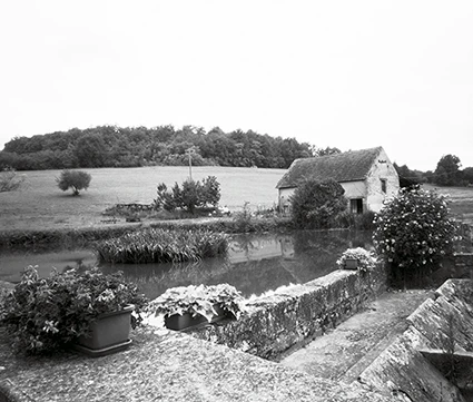 Près de la commune de Meyrals en Dordogne, sténopé d'un petit plan d'eau avec une maisonnette sur le côté et dans le lointain une petite colline boisée.
